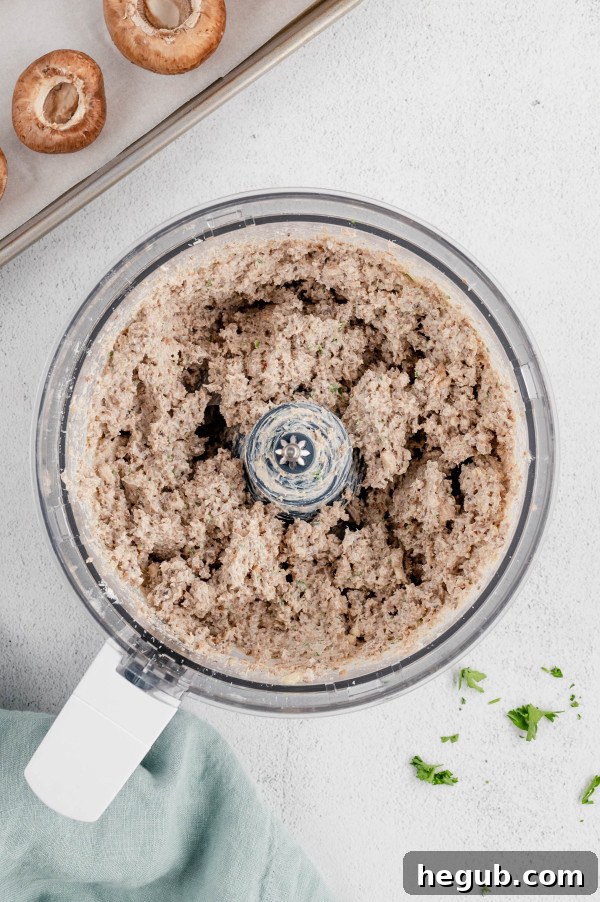 A close-up shot of a food processor bowl containing the finely chopped mixture of ground sausage, cream cheese, mushroom stems, parsley, and garlic, ready to be scooped.
