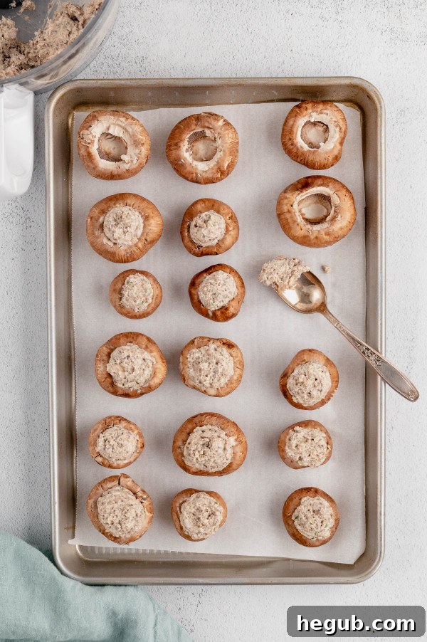 Several baby portobello mushroom caps, generously filled with the savory sausage and cream cheese stuffing, arranged neatly on a non-stick baking sheet, ready to be put into the oven.